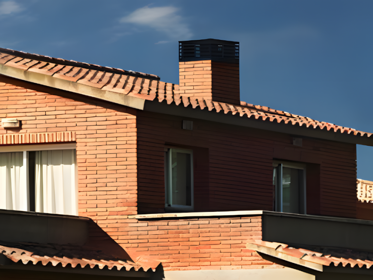 Tejado de una casa de ladrillo con chimenea y ventanas. Cielo azul al fondo.